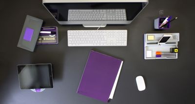 Ein dunkler Schreibtisch ist in der Draufsicht zu sehen. Dieser wurde mit verschiedenen Materialien zur Büroorganisation, wie einem Tischorganizer, Stiftehaltern und einem Tablethalter ausgestattet. Außerdem sind ein Bildschirm mit weißer Tastatur und Maus sowie ein lilafarbenes Notizbuch zu sehen.