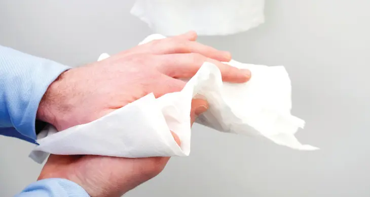 Das Bild zeigt eine Person, die sich mit einem weißen Papierhandtuch die Hände abtrocknet. Im Hintergrund ist ein Waschbecken zu sehen, und oben rechts steht der Schriftzug „Hergestellt mit Kleenex® Qualität“