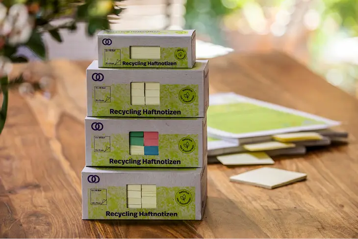 Übereinandergestapelt stehen auf einem holzfarbenem Tisch mehrere Blöcke mit Recycling-Haftnotizen der Marke oeco.