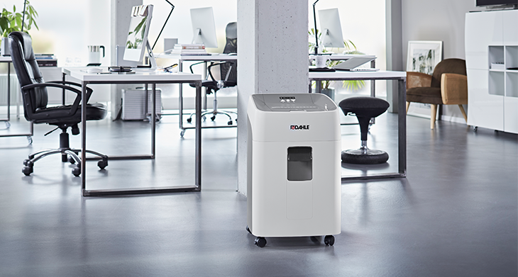 Das Bild zeigt ein modernes Büro mit einem weißen Aktenvernichter der Marke Dahle im Vordergrund. Das Büro ist mit Schreibtischen, Stühlen und minimalistischer Dekoration ausgestattet. 