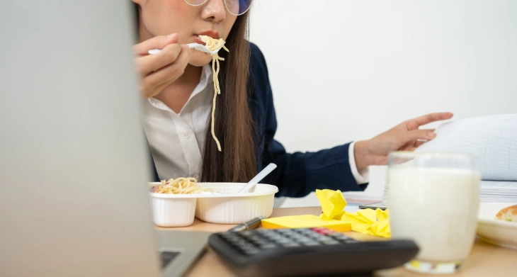 Frau isst Pasta am Schreibtisch zwischen Laptop, Unterlagen und Milchglas.