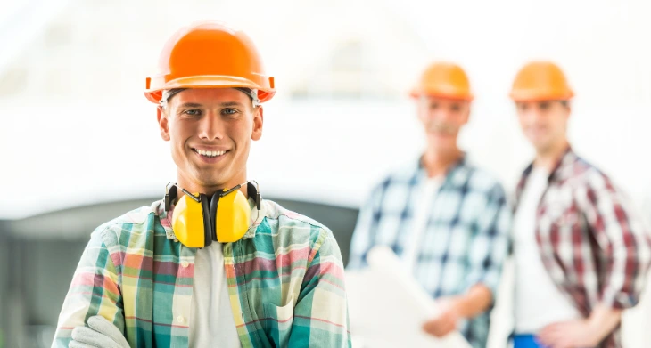 Junger Bauarbeiter mit Schutzhelm, Handschuhen und Gehörschutz lächelt, Kollegen im Hintergrund.