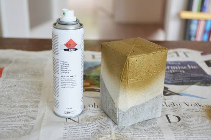 Auf Zeitungspapier ist ein Betonbecher aufgestellt, dieser wurde mit Klebeband abgeklebt und ein Teil wurde mit goldener Sprühfarbe besprüht. Die Sprühflasche steht neben dem Betonbecher.