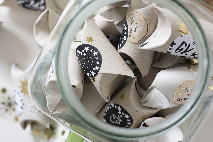 Mehrere Toilettenpapierrollen liegen als Geschenkschachtel mit einem Nummern-Aufkleber als Upcycling-Adventskalender in einem Glas.