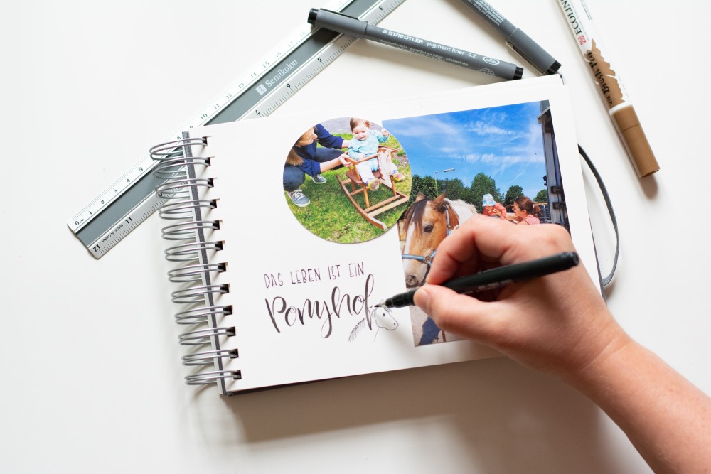Jemand lettert den Schriftzug "Das Leben ist ein Ponyhof" in ein Fotoalbum. Daneben liegen ein Lineal und Stifte.