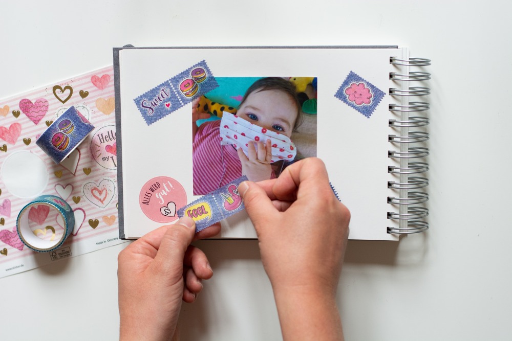 In ein Fotoalbum werden Sticker und Stücke von buntem Maskingtape eingeklebt.