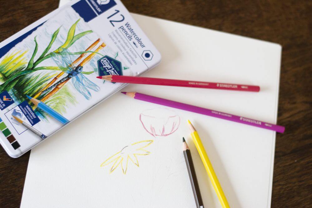 Wasservermalbare Aquarellstifte aus der Design-Journey-Kollektion von Staedtler liegen auf einem Blatt Papier. Auf das Papier sind die Umrisse eines Blumenstraußes gemalt.