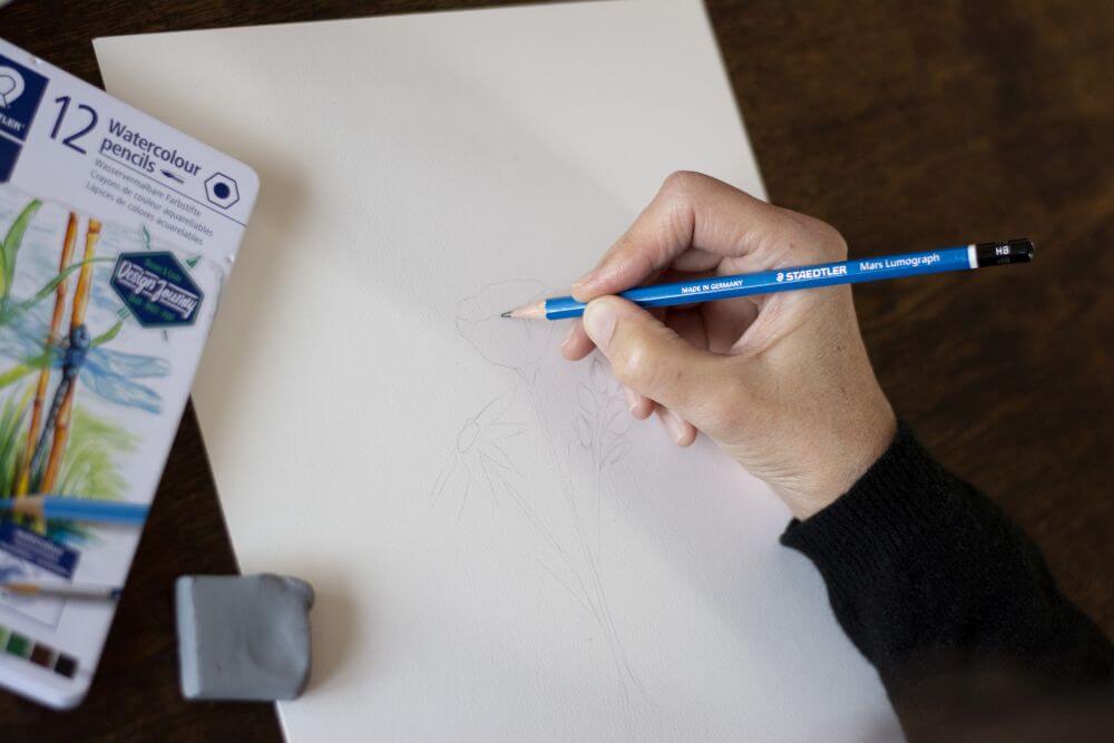 Eine Hand malt mit einem Bleistift von Staedtler sanft die Umrisse eines Blumenstraußes auf ein Blatt Papier. Daneben liegt ein knetbarer Radiergummi und eine Schachtel Aquarellstifte.