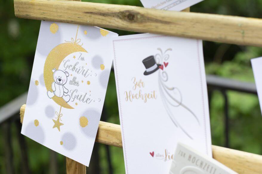 Eine Grußkarte zur Geburt und eine Glückwunschkarte zur Hochzeit hängen eingeklemmt an einem Rankgitter.