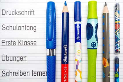 Auf einem Schreiblernblatt liegen diverse Schreiblernstifte von Pelikan: griffix Schreiblern-Tintenroller, Silbenstift, Ilo Tintenlöscher, Combino Schreiblernfüller, Combino Schreiblernbleistift sowie zwei Großraum-Tintenpatronen. Auf der linken Seite stehen verschiedene Wörter in Druckschrift. Foto: Johanna Rundel 