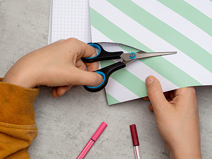 Ein Kind schneidet mit einer speziellen Linkshänder Schere von Stabilo etwas mit Links aus einem türkis/weiß gestreiften Bogen Papier aus. Foto: Johanna Rundel