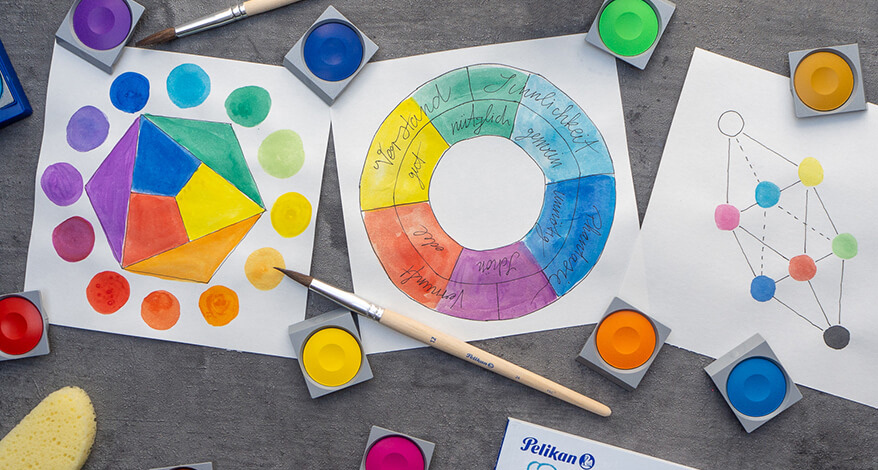 Auf einem dunkelgrauen Untergrund liegen Blätter mit den Farbkreismodellen von Itten, Goethe und Küppers. Drumherum sind bunte Farbnäpfe eines Deckfarbkastens von Pelikan verteilt. Außerdem Pinsel und Malschwämmchen. Unten rechts ragt ein Malblock ins Bild. Foto: Johanna Rundel