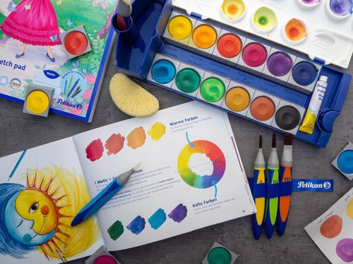 Auf einem dunkelgrauen Untergrund liegt ein aufgeklapptes Buch, in dem der Farbkreis erklärt wird. Drumherum sind verschiedene Malutensilien von Pelikan drapiert: Deckweiß, Malblock, Malschwamm, griffix-Pinsel, Wasserbehälter, Deckfarbkasten und Mischpalette. Foto: Johanna Rundel