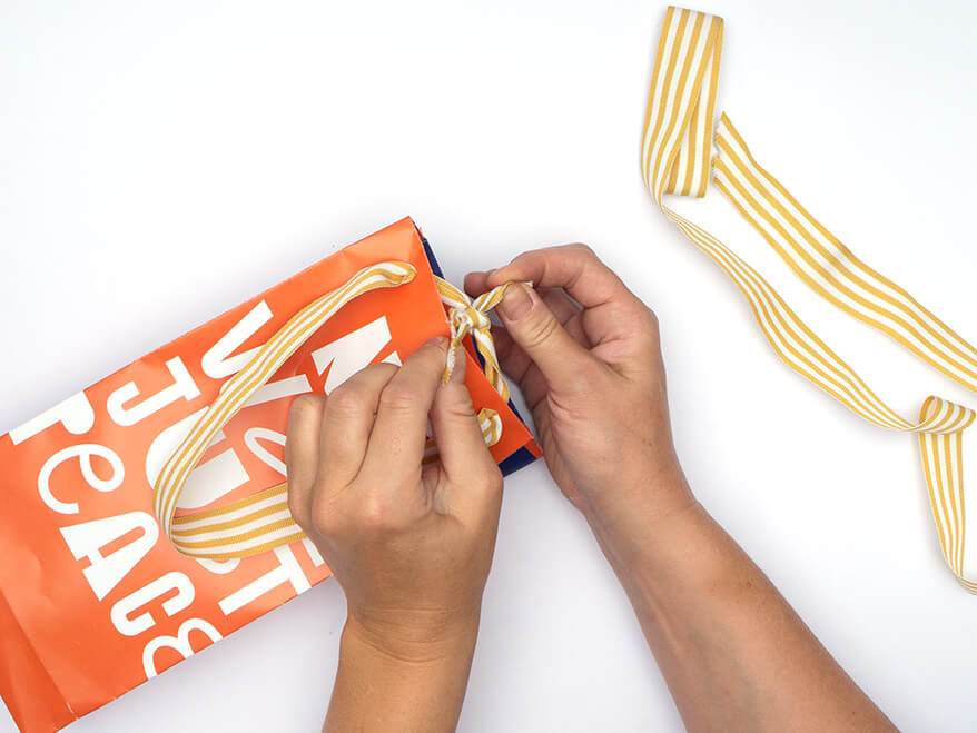 Tüte aus Papier falten ohne Vorlage: Jemand befestigt Henkel aus Stoffband an einer, aus einem rechteckigen, orangefarbenen Kalenderblatt von Athesia gefalteten, Papiertüte auseinander. Der Untergrund ist weiß. Foto: Johanna Rundel