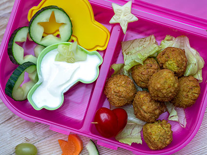 Auf einem Holzbrett liegt eine türkise Brotdose aus Plastik. In der einen Hälfte der Dose befindet sich eine kleine Plastikaufbewahrungsdose in Autoform, die mit Joghurt gefüllt ist. Außerdem sind in diesem Fach Gurkenscheiben, aus denen Sternen ausgestochen wurden. Im rechten Fach befinden sich Salatblätter, auf denen Falafelbällchen liegen. Foto: Johanna Rundel