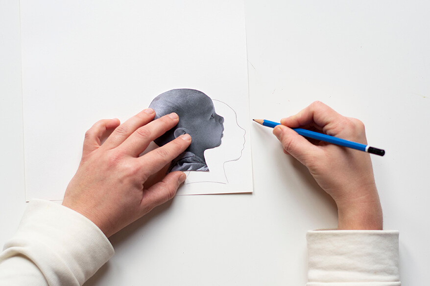 Anleitung für einen Do-it-yourself-Scherenschnitt. Schritt 2: Das ausgedruckte und ausgeschnittene Profilbild des Kindes wird auf ein Blatt cremeweißes Tonpapier gelegt und die Umrisse mit einem Bleistift umfahren.