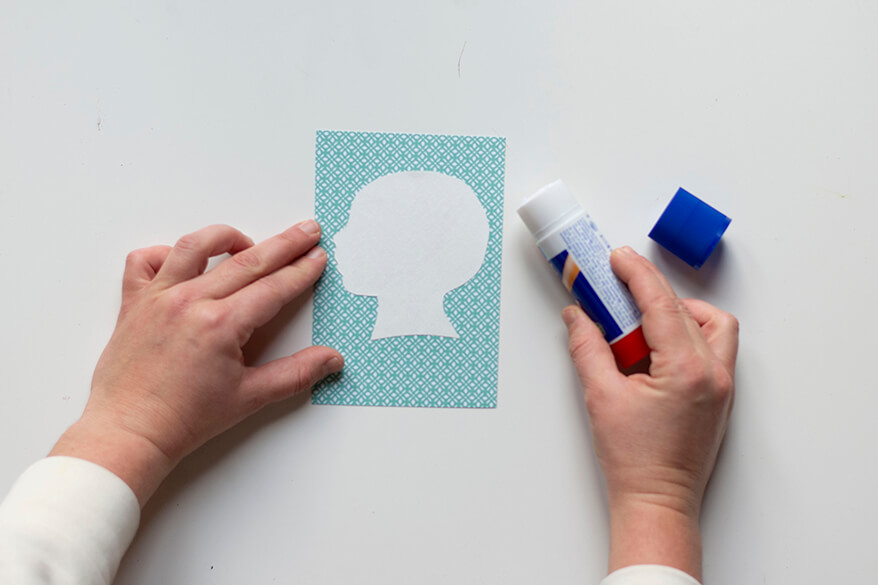 Anleitung für einen Do-it-yourself-Scherenschnitt. Schritt 5: Der cremeweiße Scherenschnitt des Kopfes des Kindes wird auf das mintfarben gemusterte Hintergrundpapier geklebt.