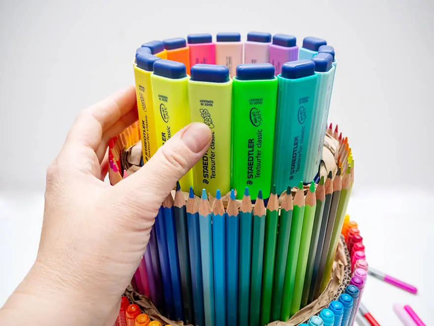 Jemand stapelt drei mit Stiften von Staedtler beklebte, immer kleiner werdende Tortenböden zu einer Stifte-Torte aufeinander.