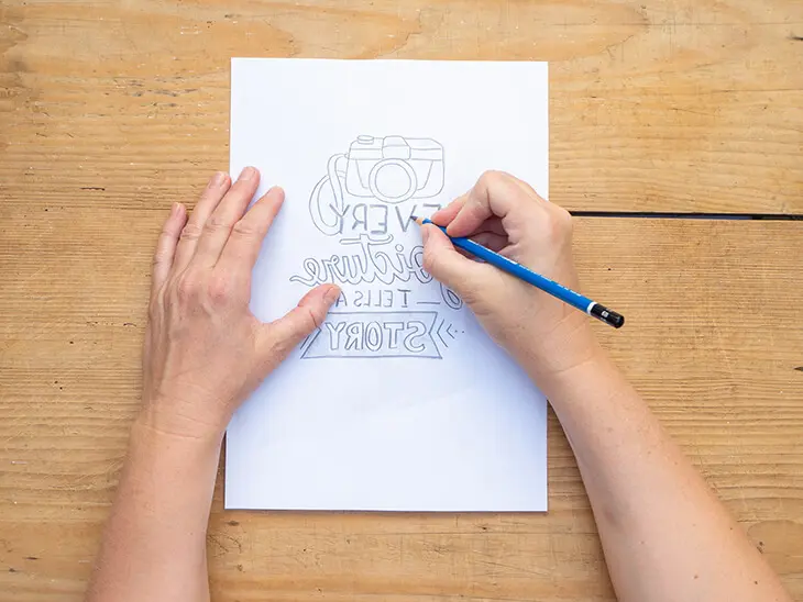 Jemand malt auf einem Holztisch eine spiegelverkehrte Letteringvorlage „Every picture tells a story“ mit einem Bleistift nach. Foto: Johanna Rundel