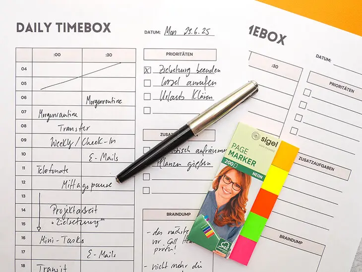 Ein ausgefüllter und ein leerer Ausdruck einer kostenlosen Downloadvorlage für ein „Daily Timeboxing“-Arbeitsblatt liegen nebeneinander auf einer gelben Schreibtischunterlage von Sigel. Foto: Johanna Rundel