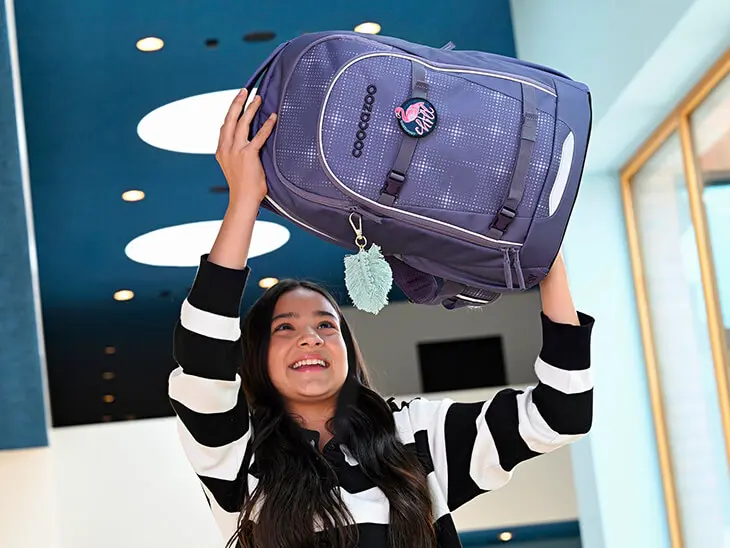 Ein Mädchen wirft seinen lilafarbenen Schulrucksack von coocazoo in die Luft.