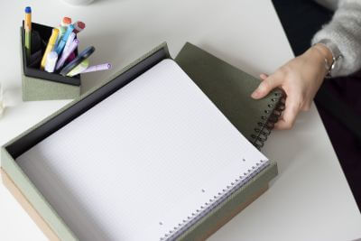 Eine Draufsicht auf einen Schreibtisch. Auf dem Schreibtisch steht ein Stifteköcher und Briefablagen der S.O.H.O. Kollektion von Rössler. Eine Hand hält ein Notizbuch, welches gerade in ein Fach der Briefablage gesteckt wird.