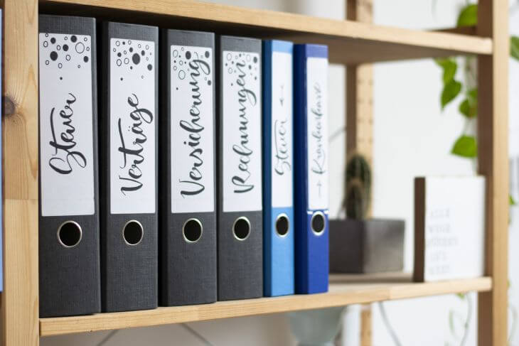 In einem Holzregal stehen vier breite, schwarze Ordner und zwei schmale Ordner in blau. Alle Ordner sind mit geletterten Ordnerrückenetiketten ausgetattet, auf den zum Beispiel "Steuer", "Veträge" und "Versicherung" steht.
