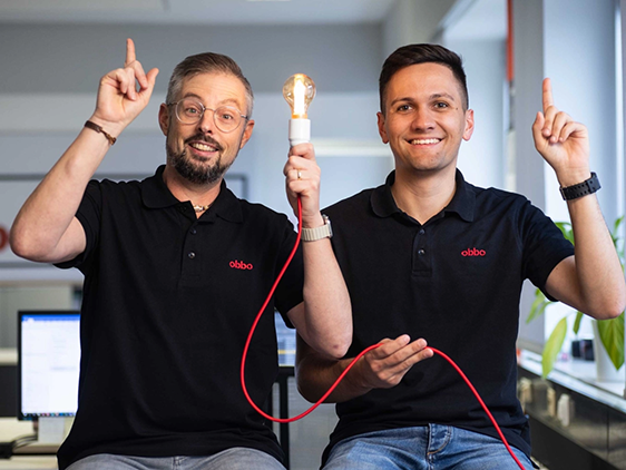 2 Männer schauen glücklich in die Kamera und halten eine Lampe in der Hand, die leuchtet