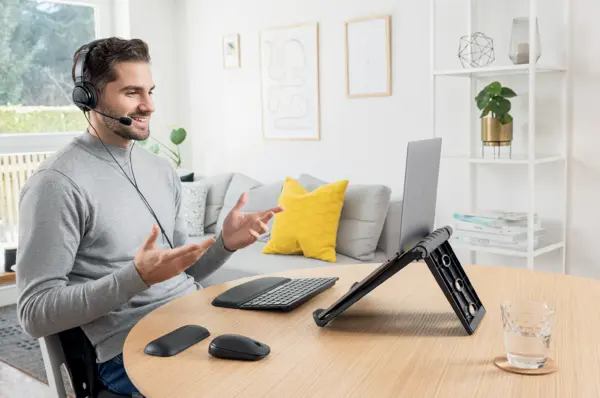 Das Bild zeigt einen Mann, der an einem Tisch mit einem höhenverstellbaren Laptopständer, externer Tastatur und Maus sitzt. Er trägt ein Headset und führt eine Videokonferenz durch.