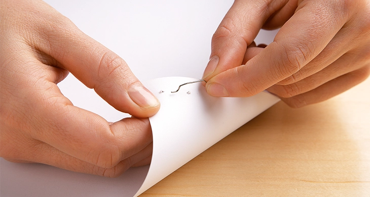 Auf dem Bild sieht man zwei Hände, die vorsichtig eine Heftklammer aus mehreren Blättern Papier entfernen. Die Person hebt das Papier leicht an, um die Klammer herauszuziehen.