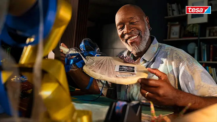 Ein lächelnder älterer Mann, der ein gerahmtes Kunstwerk an einem Basteltisch präsentiert, umgeben von tesa Klebeprodukten. Der Text lautet „Die Freude am Gelingen. tesa tadaa!