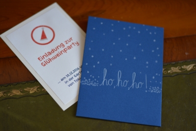 2 selbstgestaltete Karten liegen auf einem Tische. Auf einer blauen Karte steht "ho ho ho" mit Schneeflocken, auf einer weißen Karte steht "Einladung zur Glühweinparty".