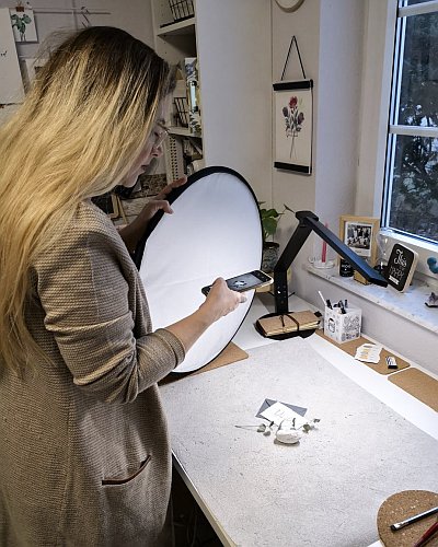 Jennifer Hertel steht vor einem Schreibtisch, auf dem eine kleine beletterte Karte und ein Zweig liegt. In der rechtebn Hand hält sie ihr Smartphone, um ein Foto zu machen, in der linken Hand hält sie eine Reflektor für eine bessere Ausleuchtung.