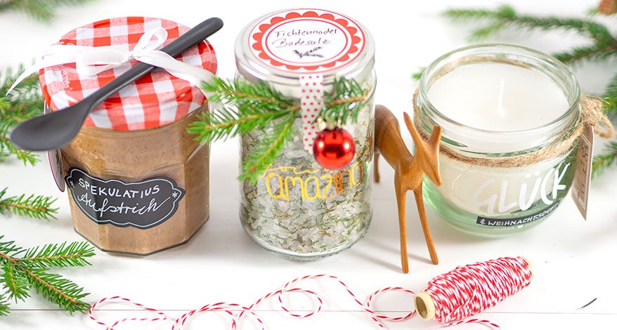 Drei schön dekorierte Gläser, gefüllt mit Spekulatius-Creme, Badesalz und einer Kreze, stehen auf einem Tisch. Der Tisch ist weihnachtlich dekoriert mit Tannenzweigen.