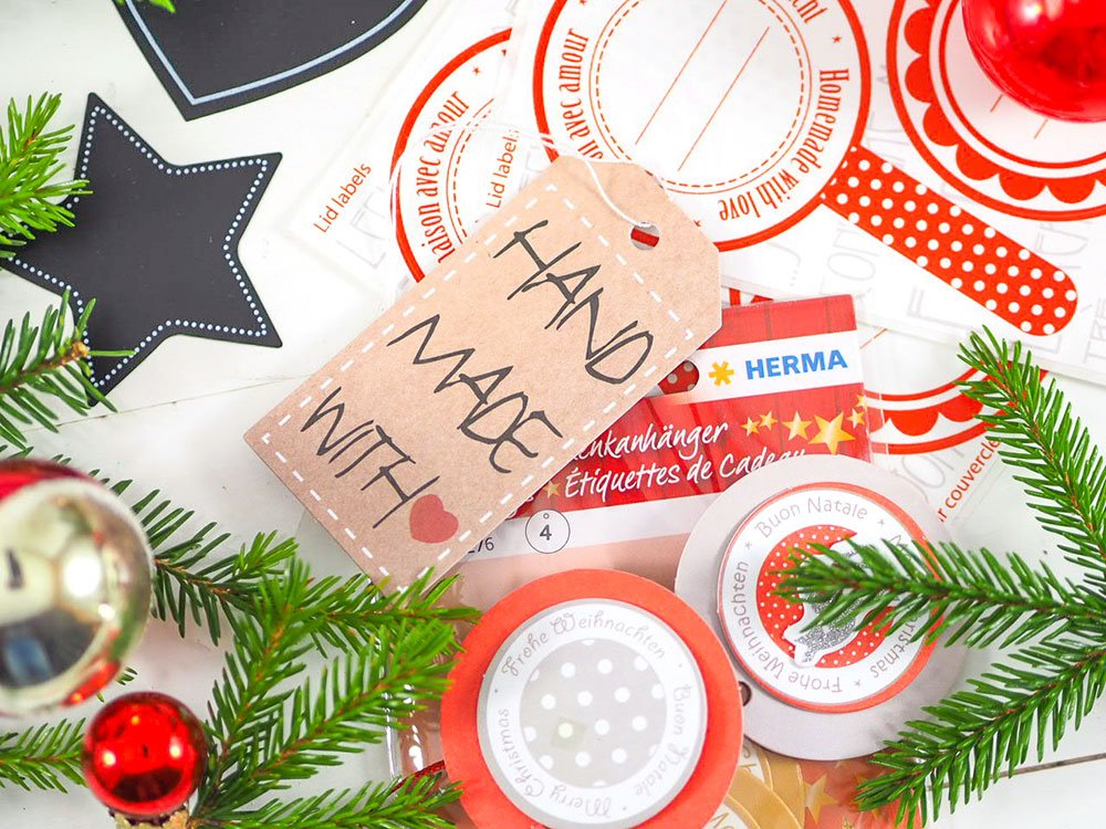 Ein Etikett der Marke HERMA mit der Aufschrift "Handmade with love" liegt auf weiteren Etiketten. Drumherum sind Tannenzweige und kleine Weihnachtskugeln dekoriert.