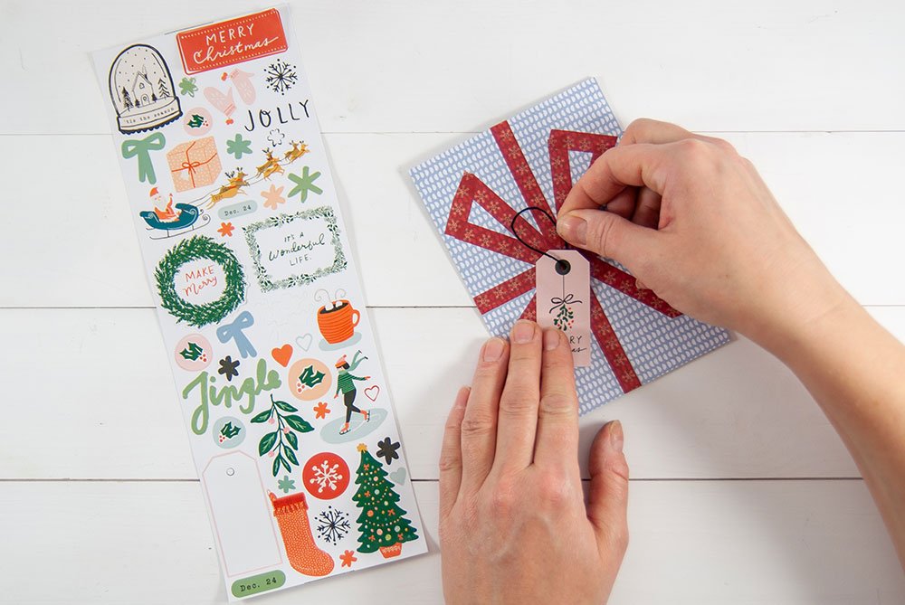 An eine selbstgebastelte Karte aus Motivkarton und Washitape, die aussieht wie ein Geschenk, wird ein Klebeetikett mit Garn angebracht. Dieses stammt von einem weihnachtlichen Stickerbogen der Marke Artoz.