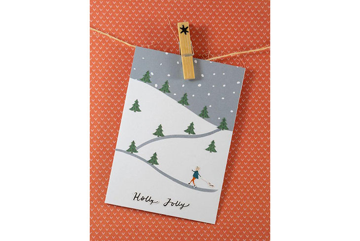 Eine grau-weiße Karte mit Wintermotiv hängt mit Hilfe einer Holzklammer an einem Garn. Der Untergrund ist rot mit weißen Punkten. Die Karte zeigt eine verschneite Winterlandschaft, in der eine Frau mit ihrem Hund spazieren geht. Auf die Karte ist „Holly Jolly“ gelettert.