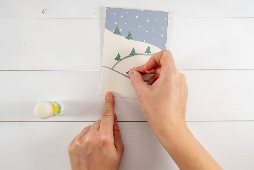 Auf eine selbstgebastelte Karte, die einen mit Schnee bedeckten Hang zeigt, werden Tannenbäume aufgeklebt. Eine Hand beklebt die Karte mit grünen Bäumen, die zuvor mit einem Motivstanzer ausgestanzt wurden. In der linken Bildecke steht ein Klebestift.