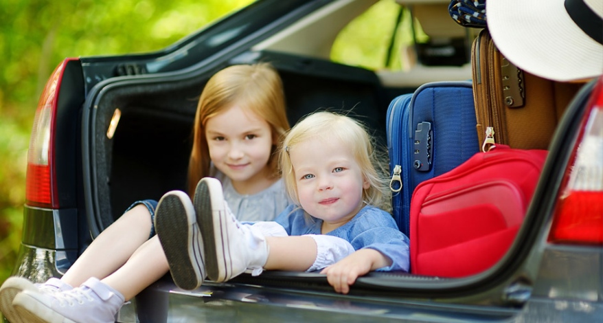 Zwei Kinder sitzen fröhlich im offenen Kofferraum eines gepackten Autos.