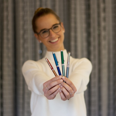 Eine junge Frau hält 4 Kugelschreiber lächelnd in die Kamera.