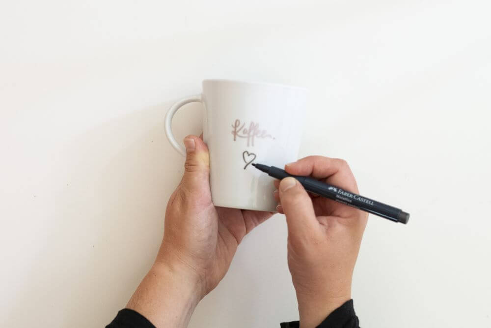 Zwei Hände halten eine weiße Tasse. Auf dieser ist ein Lettering und eine Zeichnung zu erkenne. Die Zeichnung mit einem Metallicpen von Faber Castell nachgezeichnet.