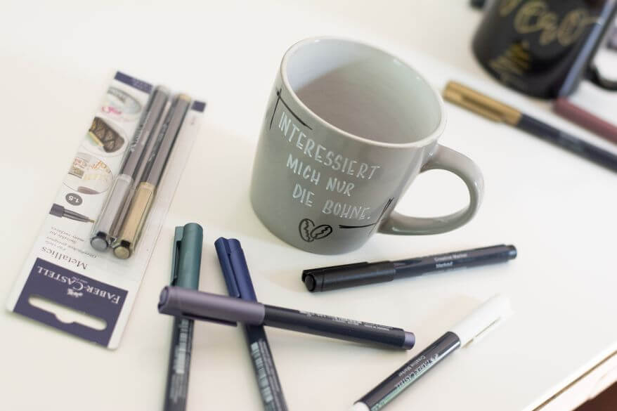Auf einem weißen Tisch steht eine graue Tasse. Um diese herum liegen verschiedene Metallic Marker von Faber Castell. Marker in silber und gold sind noch in der Produktverpackung. Die Tasse wurde mit den Metallic Markern belettert. Darauf steht: "Interessiert mich nicht die Bohne." Unter das Lettering wurden zwei Kaffeebohnen gemalt. Im Hinterrgrund sind unscharf weitere Marker und eine beletterte Tasse zu sehen.