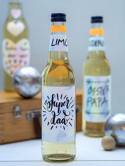 Vor einer hellblauen Wand liegt auf einem hellen Untergrund ein Boule-Koffer aus Holz. Darauf und davor stehen Limonadenflaschen mit selbstgestalteten Etiketten von Avery Zweckform mit Sprüchen zum Vatertag. Ein paar Boule-Kugeln sind zwischen den Flaschen verteilt. Foto: Johanna Rundel