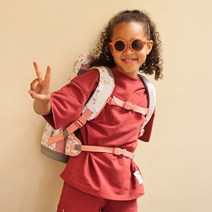 Ein Mädchen im Grundschulalter steht vor einer beigen Wand. Sie trägt eine coole Sonnenbrille, ein pinkes T-Shirt sowie eine pinke Sporthose und hat einen Schulranzen der Marke School-Mood auf dem Rücken. Das Mädchen mit den langen dunklen Locken streckt den rechten Arm nach vorne und zeigt mit ihren Fingern das Victory-Zeichen.