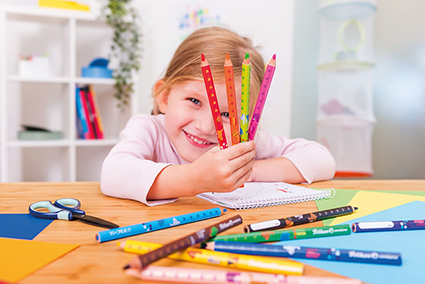 Ein blondes Mädchen im Grundschulalter hält in ihrer rechten Hand vier Buntstifte von Pelikan vor ihr Gesicht. Sie lächelt zwischen den Stiften hindurch. Vor ihr auf dem Tisch liegen weitere Bundstifte, farbige Blätter und eine Schere. Im Hintergrund ist ein hell eingerichtetes Kinderzimmer zu sehen.