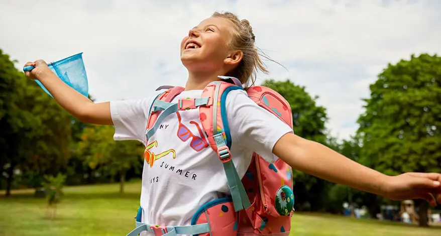 Ein blondes Mädchen im Grundschulalter hüpft fröhlich über eine Wiese in einem grünen Park. Sie trägt ein buntes T-Shirt und einen Schulranzen von ergobag. Sie blickt in den Himmel und lacht.