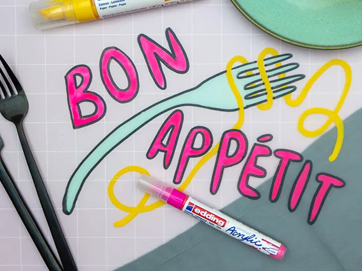 Ein durchsichtiges Tischset aus Plastik, auf das mit Acrylmarkern von edding „Bon Appétit“ sowie eine Gabel mit Spaghetti gemalt wurde, liegt auf einer rosa-karierten Tischdecke. Foto: Johanna Rundel