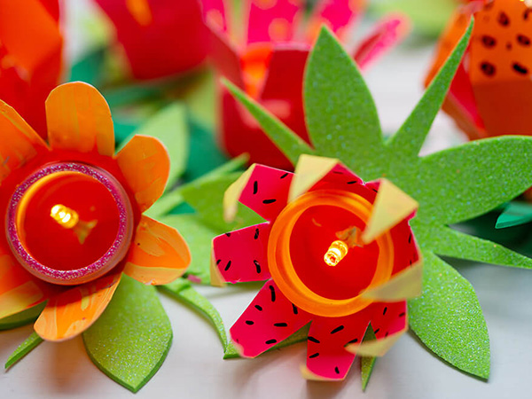 Auf einem hellen Untergrund liegt eine Lichterkette aus upcycelten Plastikflaschen in Blumenform. Die Farben der Blüten sind Orange, Pink, Rosa und Gelb. Unter den Blüten sind grüne Blätter aus Moosgummi. Die Glühbirnchen der Lichterkette leuchten. Foto: Johanna Rundel