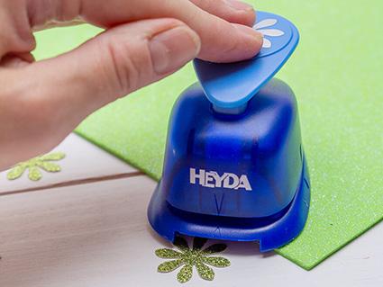 Auf einem hellen Untergrund liegt ein Bogen grünes Glitzer-Moosgummi von Knorr Prandell. Jemand stanzt daraus mit einem Stanzer von Heyda kleine Blümchen aus. Foto: Johanna Rundel