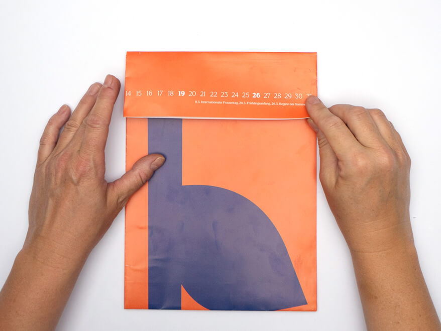 Tüte aus Papier falten ohne Vorlage: Jemand klappt ein rechteckiges, orangefarbenes Kalenderblatt von Athesia nach unten. Der Untergrund ist weiß. Foto: Johanna Rundel 